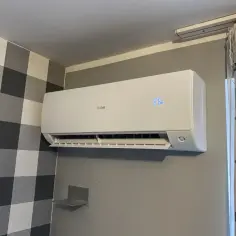 Biała klimatyzacja Haier zamontowana na szarej ścianie, obok kratowanej tapety i zasłon.