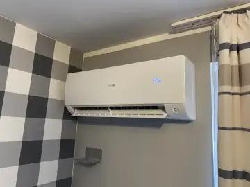 Biała klimatyzacja Haier zamontowana na szarej ścianie, obok kratowanej tapety i zasłon.